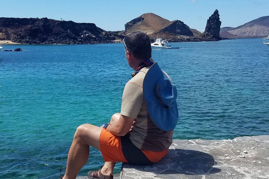 Daniel sitting on a stone ledge looking out across bright blue water toward volcanic islands